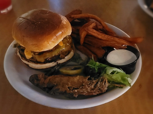 A plate with a burger topped with cheese and pickles, sweet potato fries, a side salad, and a dipping sauce.